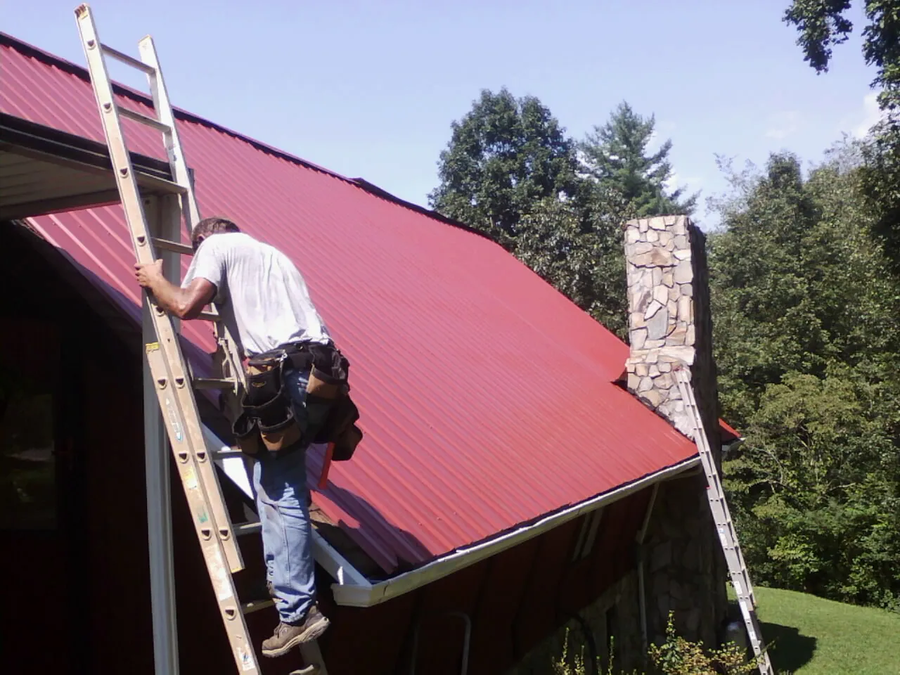 Roofer on a ladder beside a red metal roof and stone chimney — Soffit & Fascia Repair by Roofing Brothers in Putnam
